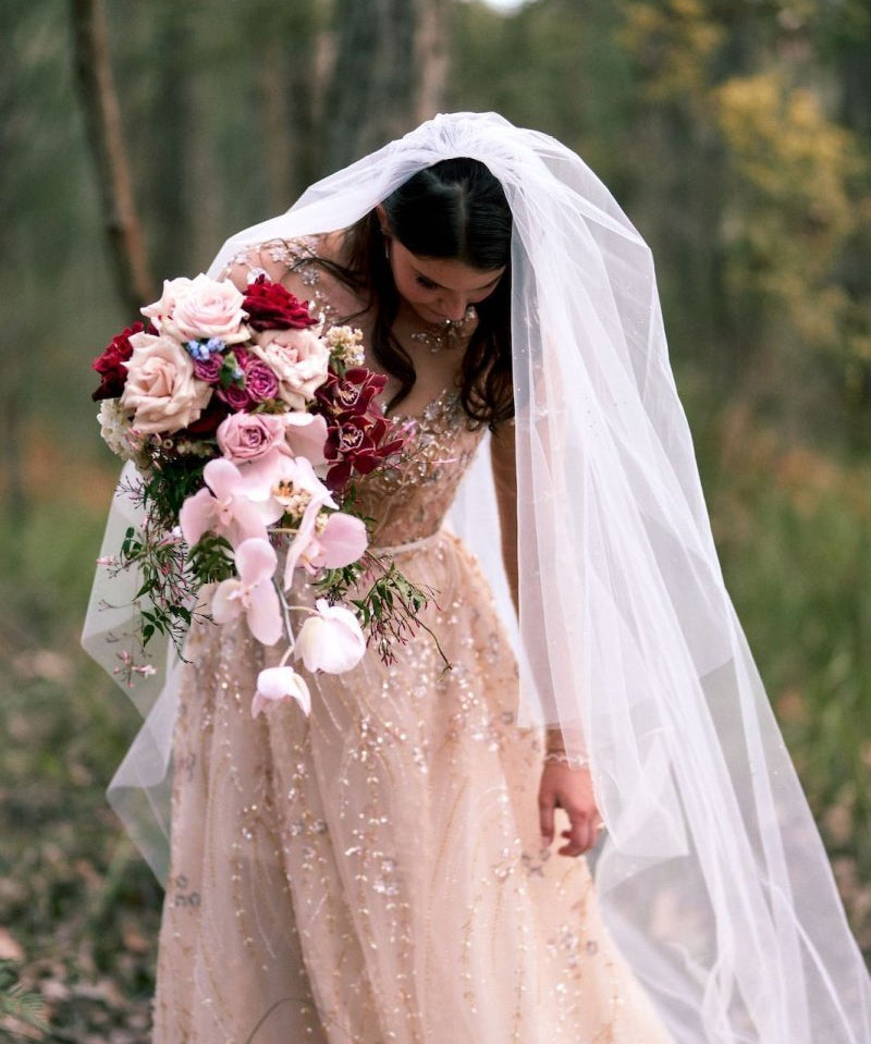 Bride in a sparkling gown holding a bouquet of flowers with a veil, standing in a natural setting.