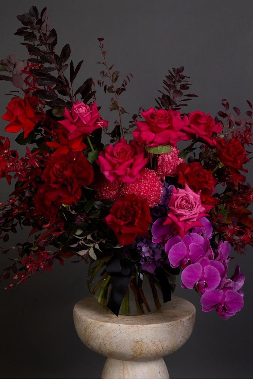 A bold bouquet Flamenco features hot-hued reflexed roses, disbuds, and luxurious phalaenopsis orchids presented with a mix of luscious contrasting foliages