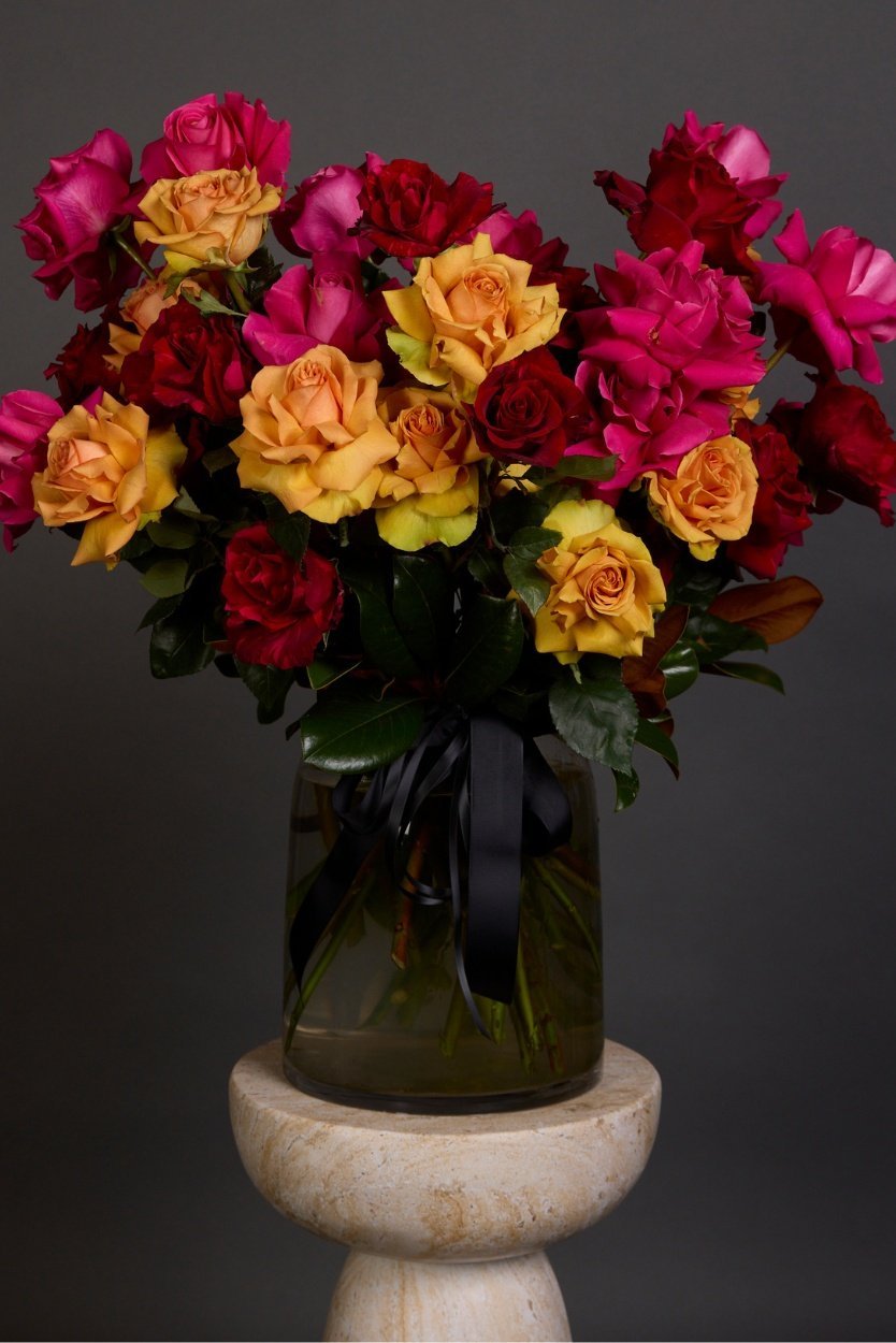 A stunningly vibrant bouquet of sunset-hued orange, pink and red long-stemmed roses styled with luscious foliage