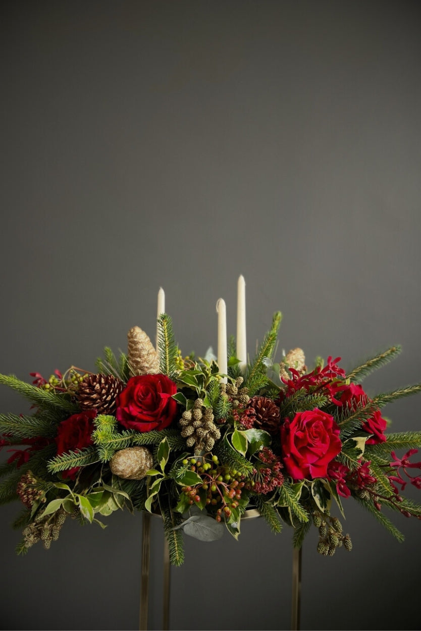 Decorative floral arrangement with red roses, greenery, and candles on a dark background