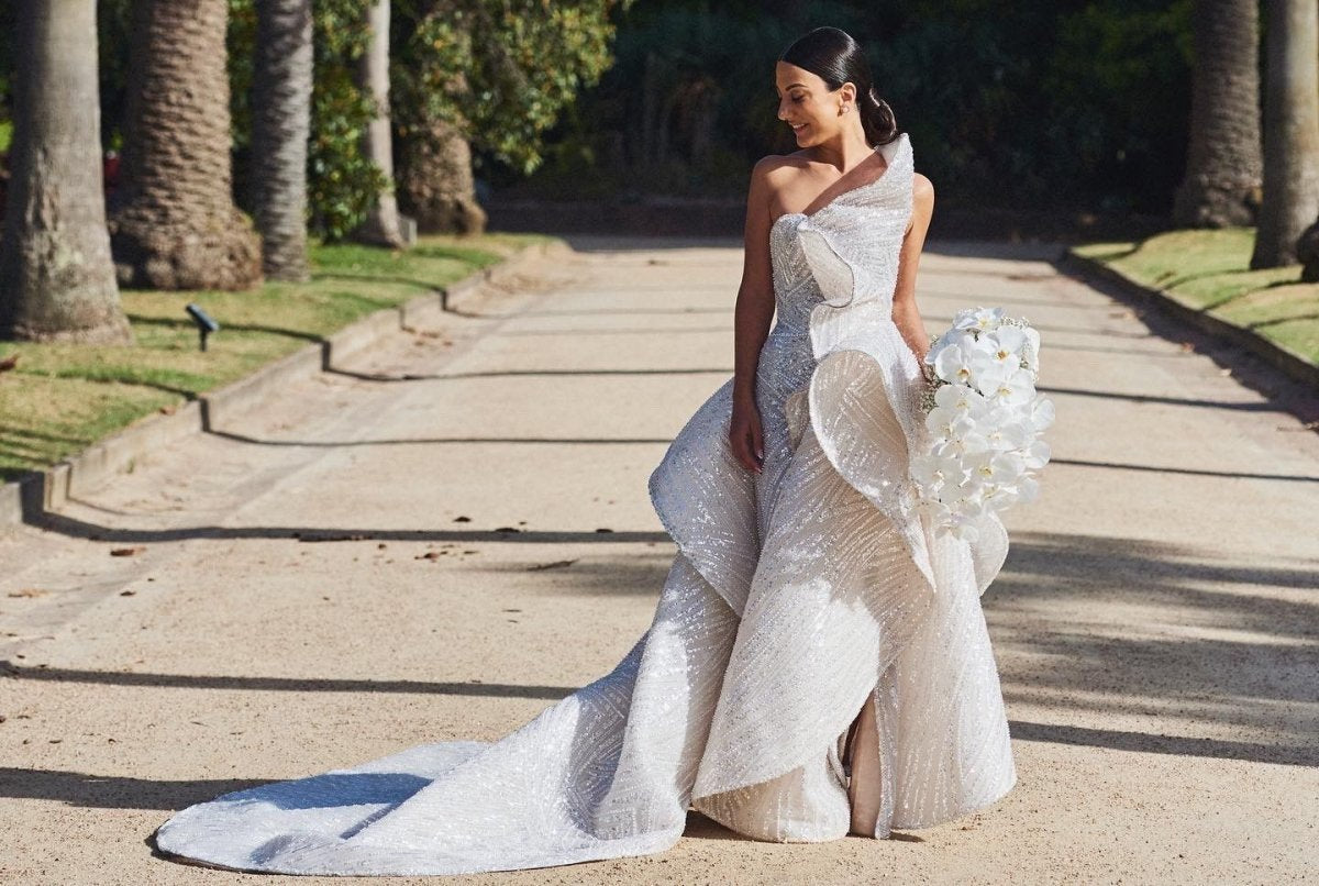 Bride in a white wedding dress holding a bouquet on a path with palm trees.
