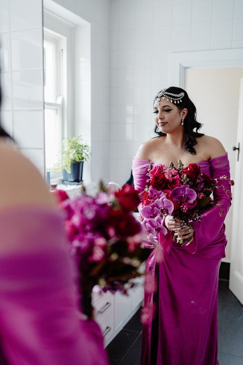 bride in a vibrant pink dress holding a bouquet of flowers in a room with white walls and a window.